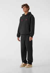 Fekete kapucnis pulóver elülső zsebbel és piros márkajellel, fekete melegítőnadrággal párosítva. Bézs sportcipővel viselve.