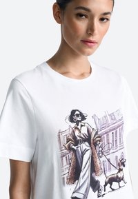 Balta kokvilnas T-krekls ar grafisku ilustrāciju, kurā attēlota sieviete, kas pastaigā suni. Dizainā iekļautas saulesbrilles un rakstaina mēteļa elements.