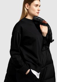 Schwarzer Pullover mit geripptem Kragen und Bündchen, der eine lockere Passform hat. Eine glänzende schwarze Pufferjacke liegt teilweise über der Schulter.
