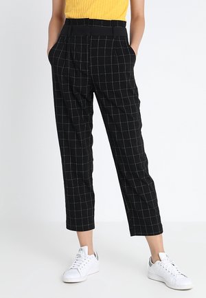 Osoba ubrana w czarne, w kratę spodnie z kieszeniami, żółtą bluzkę i białe trampki, stojąca na białym tle.