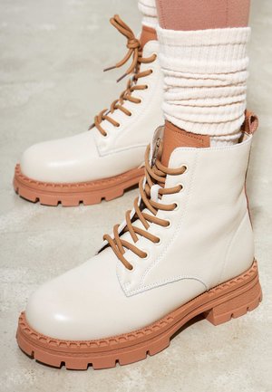 Botas de tobillo de cuero blanco con acentos marrones, que presentan una suela resistente, puntera redonda y cordones marrones, sobre un fondo neutro.