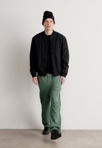 Schwarze Bomberjacke über einem schwarzen Shirt, kombiniert mit grünen Hosen und schwarzen Schuhen. Eine schwarze Mütze verdeckt kurzes Haar. Einfaches Design und lockerer Schnitt.