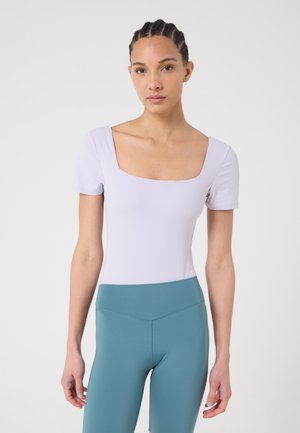 Femme aux cheveux tressés portant un haut à encolure carrée violet clair et un short ajusté taille haute couleur sarcelle, debout devant un fond uni.