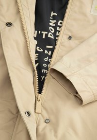 Beżowa kurtka z pełnym zamkiem, zapięciami na napy oraz czarną podszewką z białym napisem. Gładka tekstura i minimalistyczne detale wzornicze.