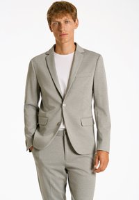 Traje gris claro a medida con un blazer de un solo botón, solapas con muesca y dos bolsillos frontales. Lleva puesto un t-shirt blanco de cuello redondo.