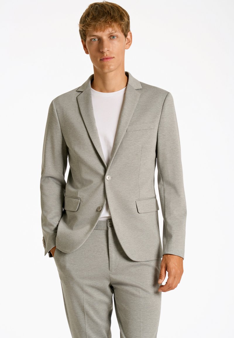 Traje gris claro a medida con un blazer de un solo botón, solapas con muesca y dos bolsillos frontales. Lleva puesto un t-shirt blanco de cuello redondo.