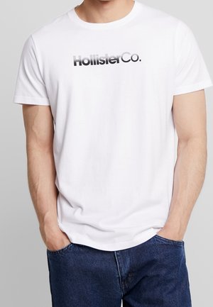 Mann trägt weißes kurzärmeliges T-Shirt mit „HollisterCo.“ Logo, die Hände in den Taschen, kombiniert mit dunkelblauen Jeans.