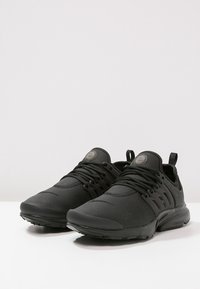 Černé sportovní boty s texturovaným povrchem, minimalistickým designem, tkaničkami, polstrovaným límcem a gumovou podrážkou pro lepší přilnavost.