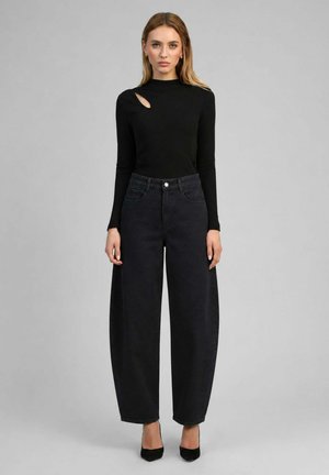 Femme debout, regardant vers l’avant, portant un haut noir à manches longues avec une découpe aux épaules, un jean noir à jambes larges et des talons noirs.