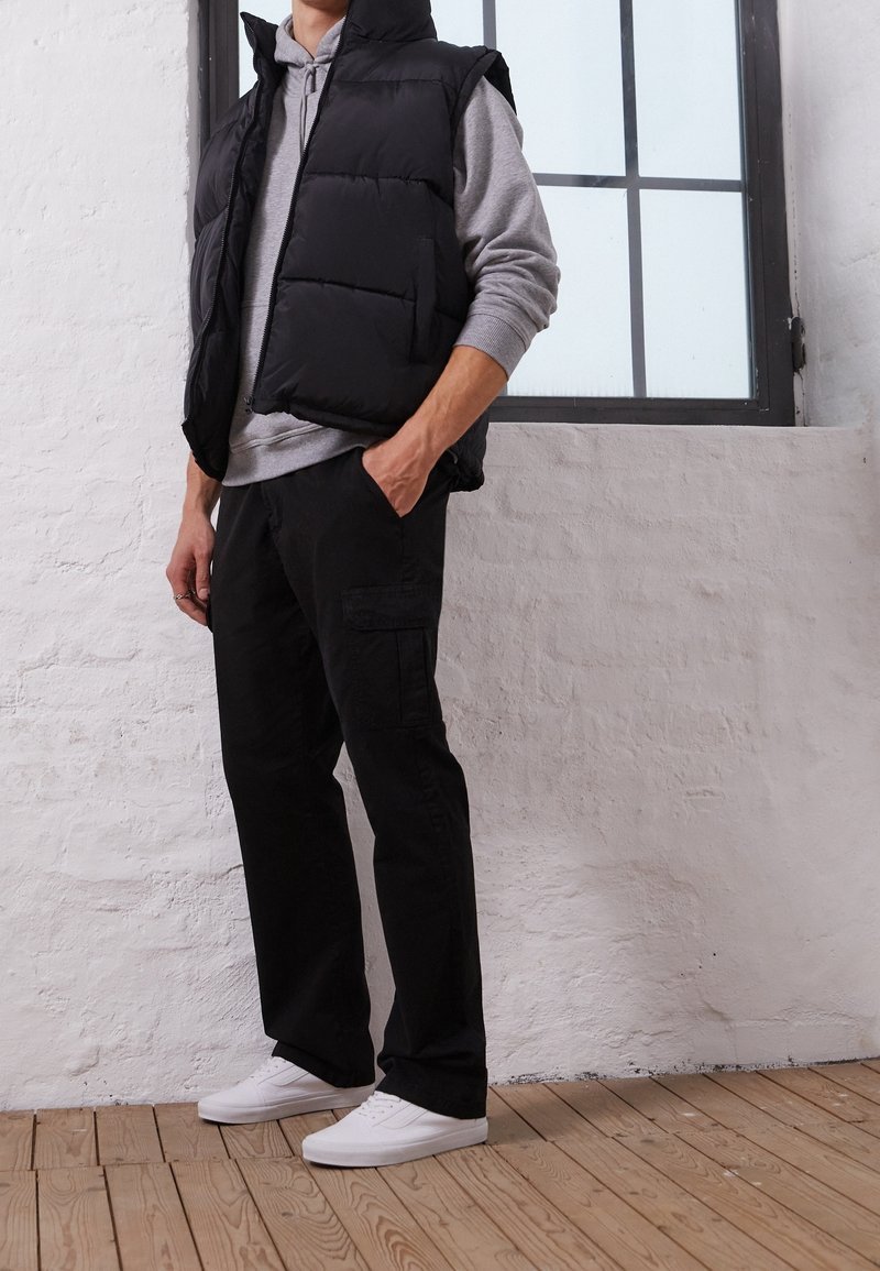 Gilet imbottito nero sopra una felpa grigia, abbinato a pantaloni cargo neri e sneakers bianche. Design semplice con texture trapuntata.