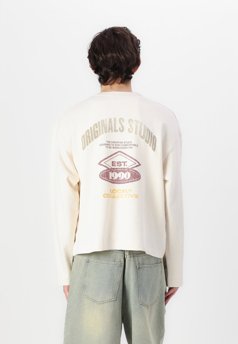 Chemise à manches longues couleur crème avec une coupe décontractée, arborant un imprimé graphique au dos en couleurs métalliques. Tissu en coton doux avec une finition légèrement texturée.