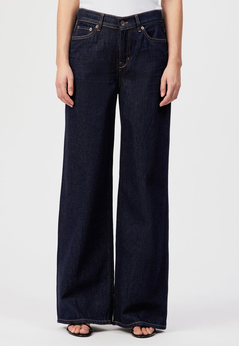 Femme portant un jean denim bleu foncé à jambes larges et des sandales noires à bout ouvert, debout devant un fond clair uni.