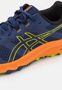Blauer Trailrunning-Schuh mit Mesh-Obermaterial, schwarzen Akzenten, gelbem Logo und orangefarbener Sohle. Verfügt über eine strukturierte Oberfläche und ein robustes Profil für besseren Halt.