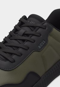 Κοντινό πλάνο ενός μαύρου και ελιάς πράσινου αθλητικού παπουτσιού BOSS με μαύρα κορδόνια και υφή στην σόλα, που δείχνει λεπτομερή ραφή και το όνομα της μάρκας στο πλάι.