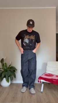 Tricou grafic negru cu imprimeu auriu, blugi dark blue și papuci gri deschis. Fundalul include o plantă verde și un scaun alb cu o pătură roșie.