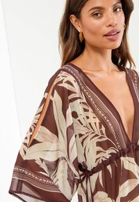 Kaftan marrone con stampa floreale, scollo a V profondo, fessure corte sulle maniche e vita arricciata. Tessuto leggero e semi-trasparente.