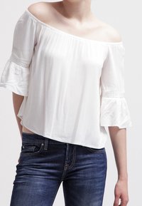 Top bianco off-the-shoulder con maniche a ruffles, dotato di dettagli in pizzo vicino ai polsini, abbinato a jeans blu scuro. Tessuto morbido e fluido.