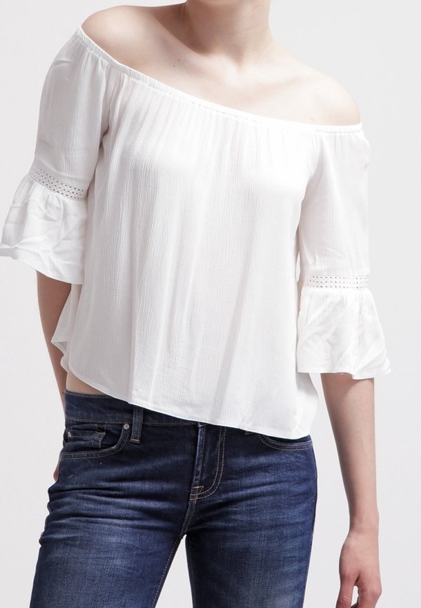 Top bianco off-the-shoulder con maniche a ruffles, dotato di dettagli in pizzo vicino ai polsini, abbinato a jeans blu scuro. Tessuto morbido e fluido.