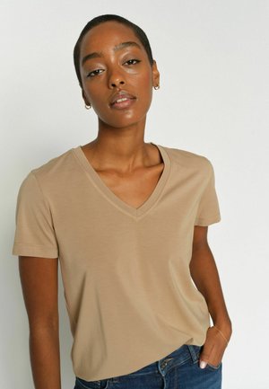 Žena s krátkými vlasy v béžovém tričku s výstřihem do V a modrých džínách, pózující s jednou rukou v kapse na jednoduchém pozadí.