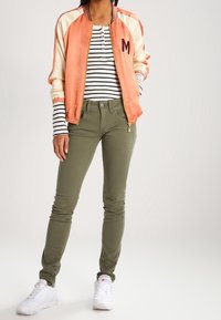 Orange och krämfärgad satin bomberjacka med ett bokstaven M, randig långärmad t-shirt, olivgröna slim-fit byxor och vita sneakers.