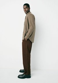 Man wearing a beige plaid shirt, brown pants, and black shoes stands in profile with hands in pockets against a white background.