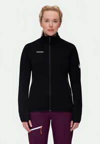 Ženska oblečena v črno jakno Mammut z zadrgo in vijolične hlače, stoji obrnjenaz naprej proti enobarvni beli ozadju.