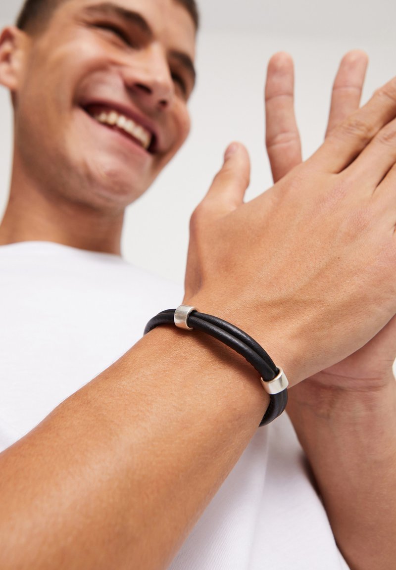Pulsera de cuero negro con detalles en plata; presenta un diseño de doble hebra que se lleva en la muñeca, mostrando una textura lisa.