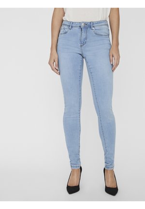 Femme portant un jean skinny bleu clair et des escarpins noirs à bouts pointus, debout contre un fond uni clair.