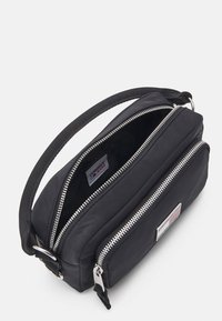 Czarna torba materiałowa o prostokątnym kształcie, z podwójnymi srebrnymi suwakami, plecioną rączką oraz naszywką z logo z przodu. Widać wewnętrzną komorę.