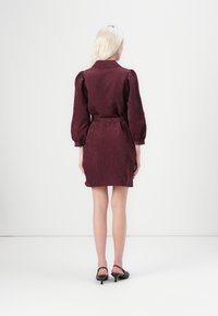 Burgunderfarbendes Cord-Kleid mit Kragen, puffärmel und gebundenem Taillenbereich. Das Kleid hat eine figurnahe Silhouette und ist knielang.
