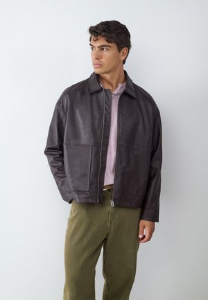 Giovane uomo che indossa una giacca di pelle marrone scuro, una camicia rosa pallido e pantaloni verde oliva, che guarda a sinistra su uno sfondo bianco semplice.