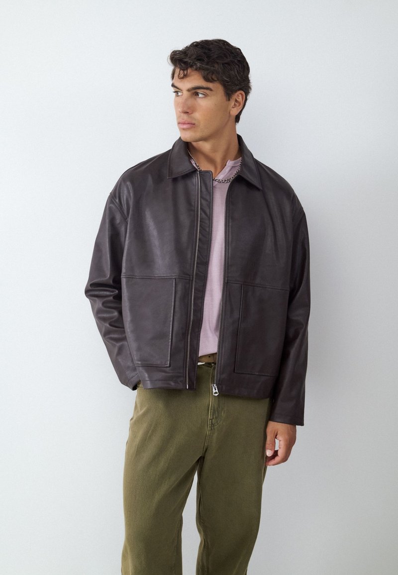 Young man wearing a dark brown leather jacket, pale pink shirt, and olive green pants, looking to his left against a plain white background.