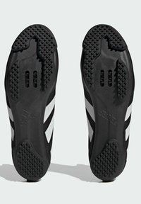 Chaussures de sport noires avec des rayures blanches, présentant une semelle texturée et deux encoches près du talon pour une meilleure adhérence et stabilité.