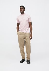 Šviesiai rožinė polo marškinėliai su apykakle ir subtiliu logotipu. Porą smėlio spalvos kelnių ir juodi batai. Švarus dizainas su lygiu paviršiumi.