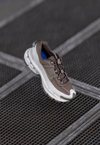 Καφέ και λευκό αθλητικό παπούτσι με υφή στο επάνω μέρος, ημι-reflective λεπτομέρειες και παχύ πάτο με ελαφριά καμπύλη. Ορατό μπλε εσωτερικό.