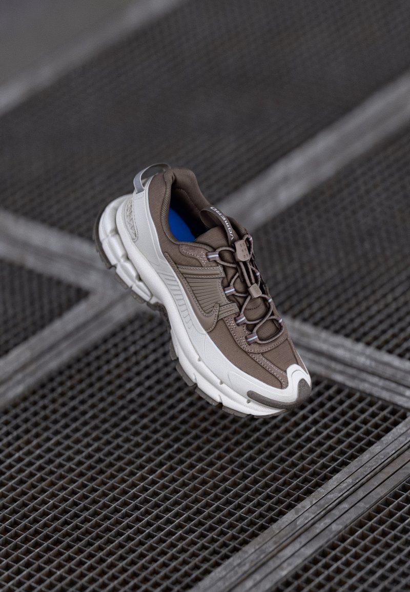 Καφέ και λευκό αθλητικό παπούτσι με υφή στο επάνω μέρος, ημι-reflective λεπτομέρειες και παχύ πάτο με ελαφριά καμπύλη. Ορατό μπλε εσωτερικό.
