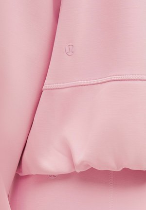 Haut à capuche rose clair en tissu lisse et élastique. Présente un petit logo et une couture visible sur l'ourlet. Texture douce et coupe décontractée.