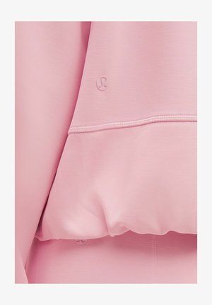 Sudadera rosa claro con tejido suave y elástico. Presenta un pequeño logo y costuras visibles en el dobladillo. Textura suave y corte relajado.