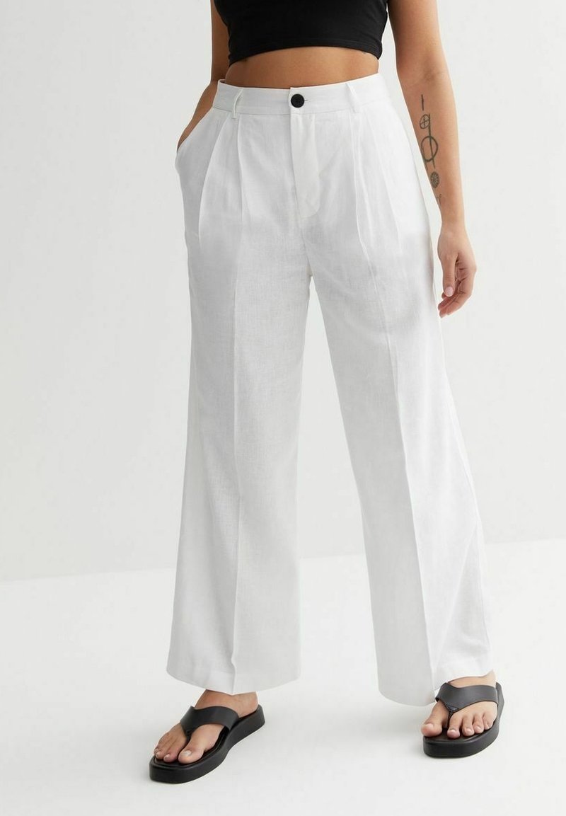 Vita linnestora byxor med framfickor, midjeknäppning och sidofickor. Bärs med svarta flip-flops och en svart kort topp.