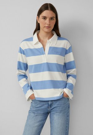 Junge Frau trägt ein langärmeliges, blau-weiß gestreiftes Rugbyshirt mit Kragen, kombiniert mit hellblauen Jeans, die Hände in den Taschen.