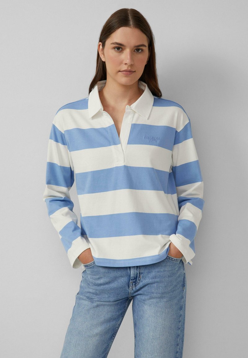 Junge Frau trägt ein langärmeliges, blau-weiß gestreiftes Rugbyshirt mit Kragen, kombiniert mit hellblauen Jeans, die Hände in den Taschen.