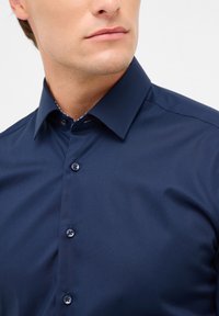 Camicia bottonata blu navy con tessuto texture, colletto pieghevole e bottoni abbinati. Un dettaglio a motivo fine è visibile all'interno del colletto.