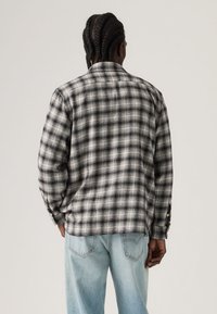 Άνδρας με πλεξούδες, φορά μακρυμάνικο πουκάμισο σε ασπρόμαυρο καρό και ανοιχτόχρωμο τζιν, στέκεται έχοντας την πλάτη του προς τα εμάς.