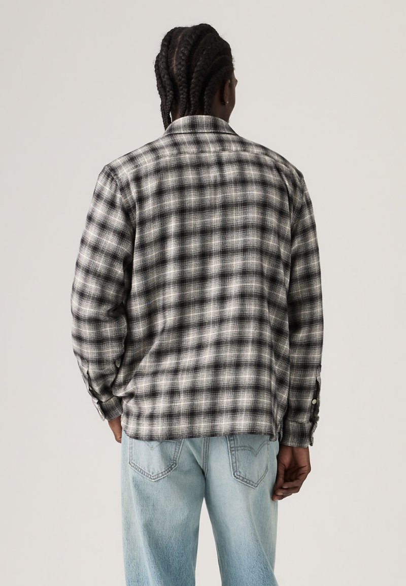 Homme aux cheveux tressés portant une chemise à carreaux noir et blanc à manches longues et un jean en denim bleu clair, debout de dos.