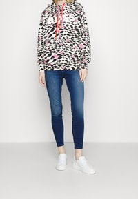Felpa colorata con stampa astratta nera, bianca e rosa, caratterizzata da un logo frontale e cordini rossi, abbinata a jeans aderenti blu e sneakers bianche.