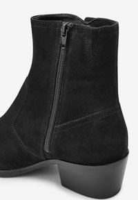 Bottine en daim noir avec fermeture éclair latérale, talon empilé et coutures nettes, avec une texture lisse et un design élégant et moderne.