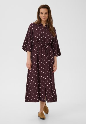 Vrouw draagt een paarse midi-jurk met lichtroze stippen, knopen aan de voorkant, tailleband en beige instapschoenen, staand op een witte achtergrond.
