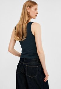 Dunkelblaues ärmelloses Oberteil mit figurnahem Schnitt, kombiniert mit hochgeschnittenen, dunklen Jeans aus Denim, die sichtbare Nähte und zwei Gesäßtaschen aufweisen.