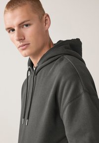 Dunkelgrauer Kapuzenpullover aus weichem Stoff, mit einer Kordelzugkapuze, lockerer Passform und sichtbaren Nähten auf den Schultern.
