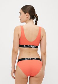 Koralinis, įribuotas bikini rinkinys, pasižymintis žemu nugaros kirpimu ir atitinkamais apatiniais, papuoštais juodu Calvin Klein logotipo juosteliu. Lygus paviršius.